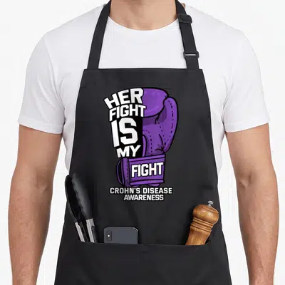 Fight crohns awareness apron - Purple cartoon boxing glove with white text HER FIGHT IS MY FIGHT CROHN'S DISEASE — shown on a black cotton canvas apron against a white background