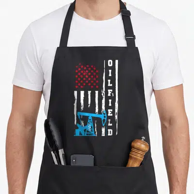 Oilman patriotic apron - Vertical OILFIELD text beside blue pump jack over red stars and white stripes — shown on a black cotton canvas apron against a white background