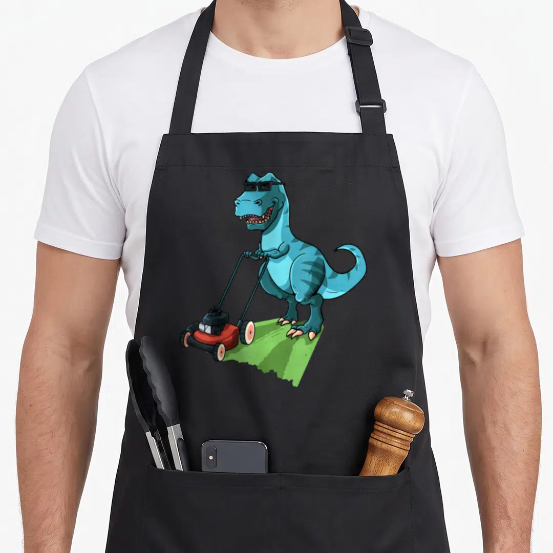 Lawn t-rex apron - Blue cartoon T-Rex wearing sunglasses pushing red lawnmower on green grass patch — shown on a black cotton canvas apron against a white background
