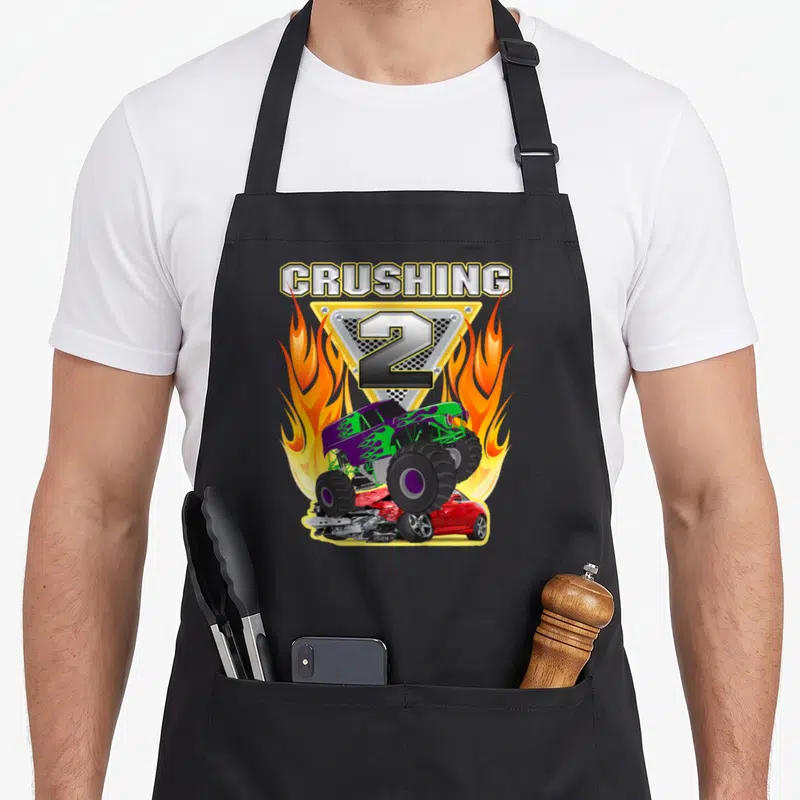 Monster crushing truck apron - CRUSHING 2 text above purple green monster truck crushing red car with orange flames — shown on a black cotton canvas apron against a white background