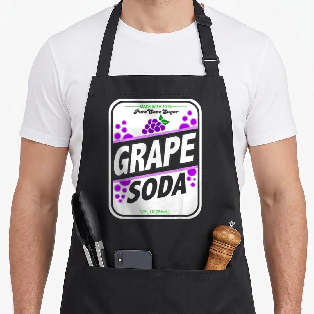Soda grapes apron - Retro soda can label print with purple bubbles, green leaves, and stacked GRAPE SODA text on white — shown on a black cotton canvas apron against a white background