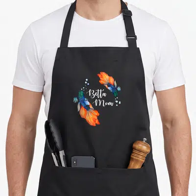Funny pet fish apron - Two blue and orange betta fish framing white Betta Mom script with scattered bubbles — shown on a black cotton canvas apron against a white background