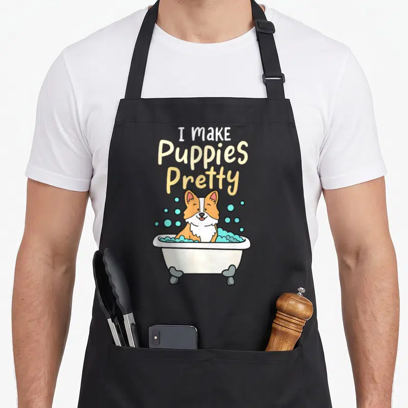 Pretty dog groomer apron - I Make Puppies Pretty text above a cartoon corgi in a blue bubble bath — shown on a black cotton canvas apron against a white background