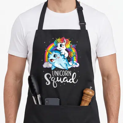 Squad rainbow apron - Cartoon unicorn riding blue narwhal under rainbow with white UNICORN Squad text — shown on a black cotton canvas apron against a white background