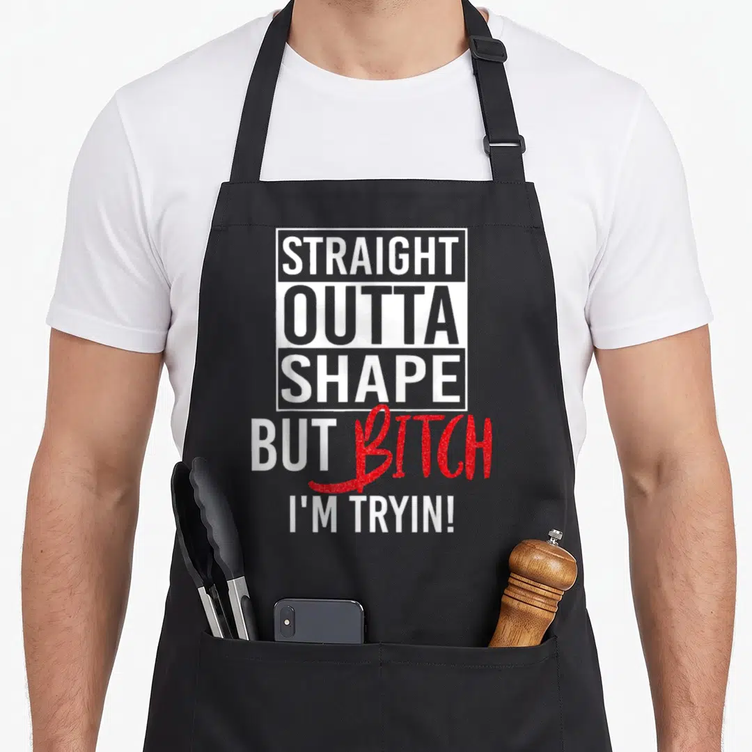 Straight shapes apron - Stacked white text in a box with red glitter script saying STRAIGHT OUTTA SHAPE BUT BITCH I'M — shown on a black cotton canvas apron against a white background