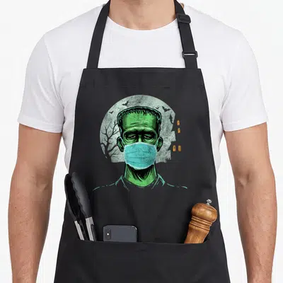 Mask frankenstein apron - Green zombie wearing blue face mask inside grey circle with tree silhouette — shown on a black cotton canvas apron against a white background