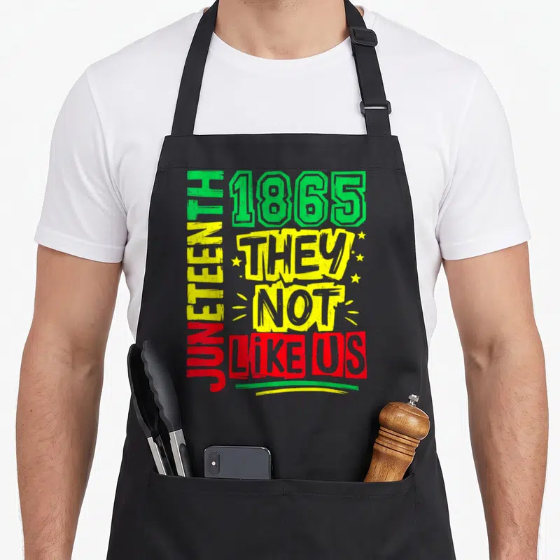 They us aprons - Text JUNETEENTH 1865 THEY NOT LIKE US in green yellow and red distressed font — shown on a black cotton canvas apron against a white background