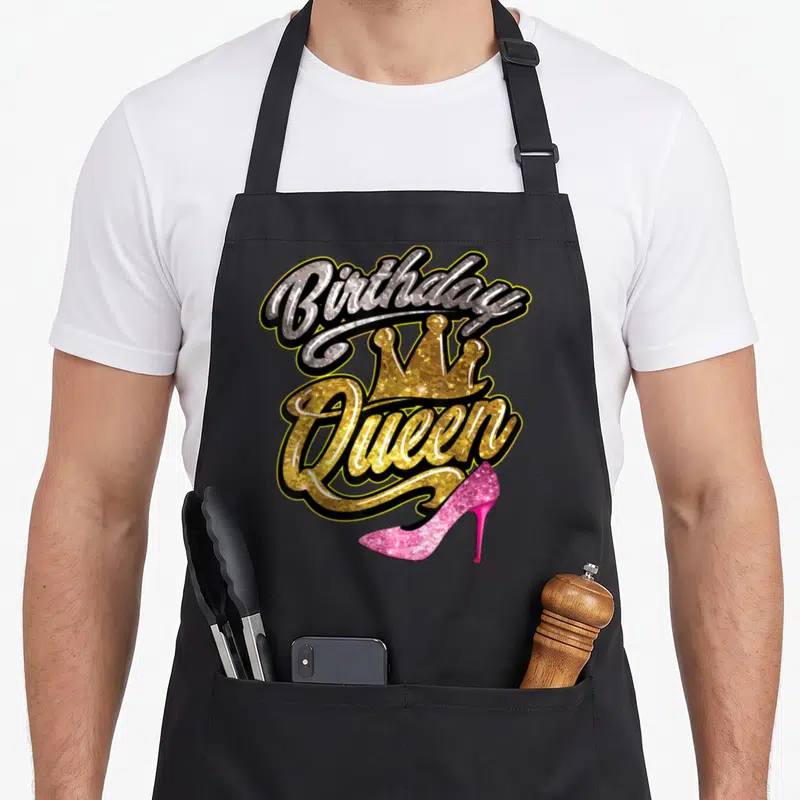 Birthday special aprons - Glittery Birthday Queen text with gold crown and pink high heel shoe graphic — shown on a black cotton canvas apron against a white background