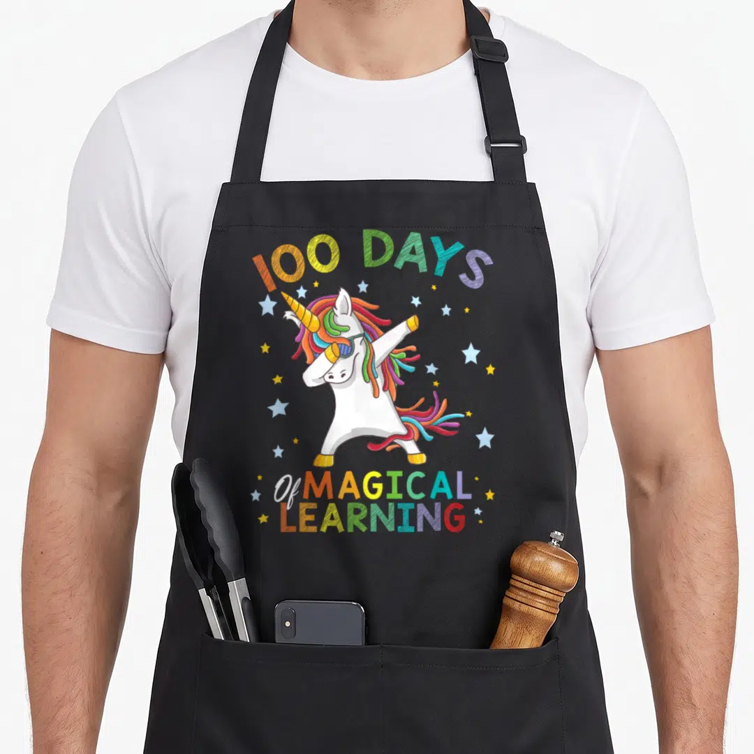 Learning unicorn apron - Dabbing white unicorn with rainbow mane and 100 DAYS of MAGICAL LEARNING text surrounded — shown on a black cotton canvas apron against a white background