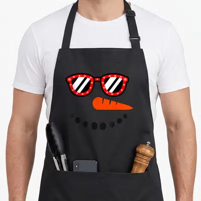 Funny christmas snowman apron - Cartoon snowman face with red snowflake glasses, orange nose, and black dot smile — shown on a black cotton canvas apron against a white background