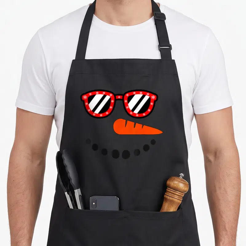 Funny christmas snowman apron - Cartoon snowman face with red snowflake glasses, orange nose, and black dot smile — shown on a black cotton canvas apron against a white background