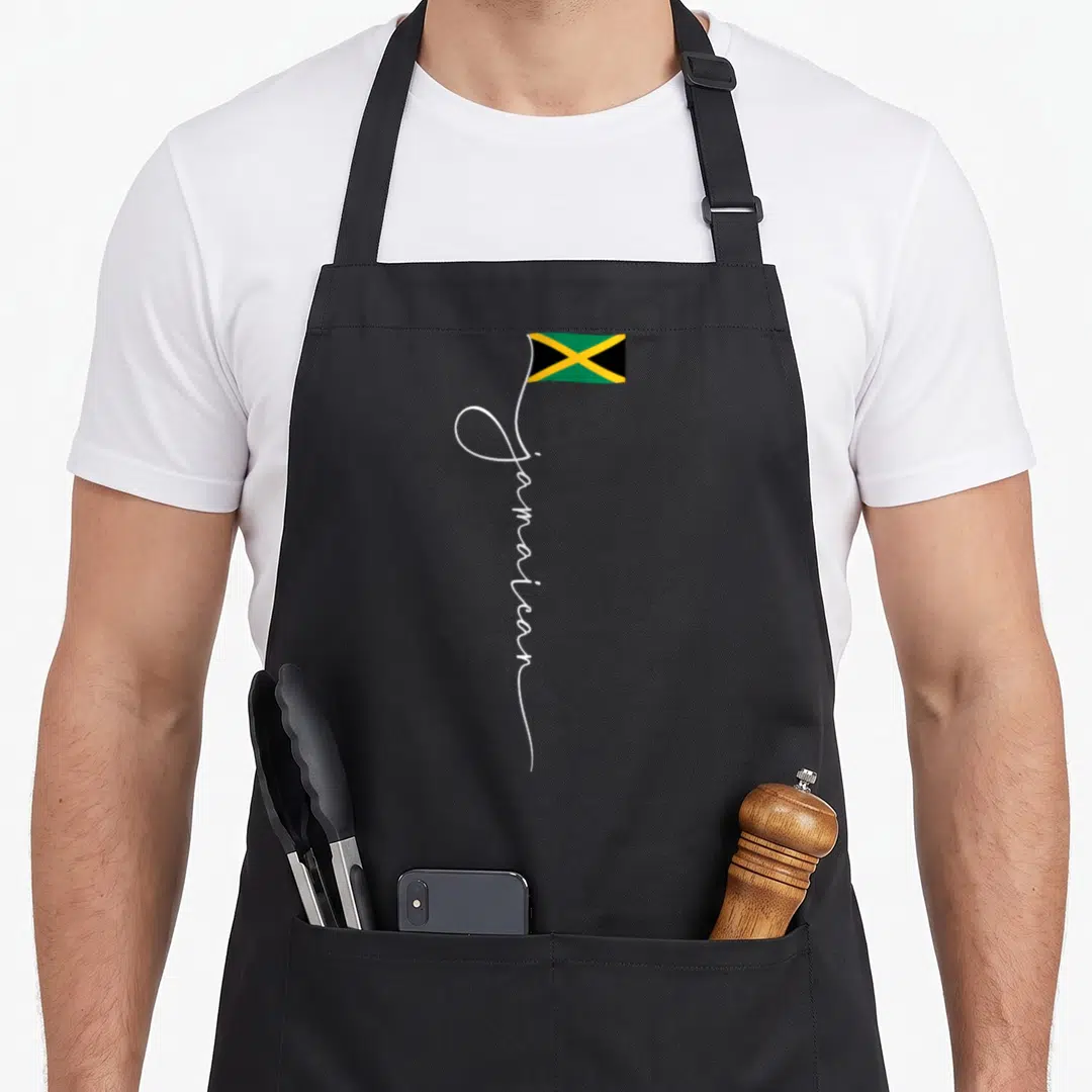 Signature jamaican apron - Vertical white script spelling Jamaican extending into a small green yellow and black flag — shown on a black cotton canvas apron against a white background