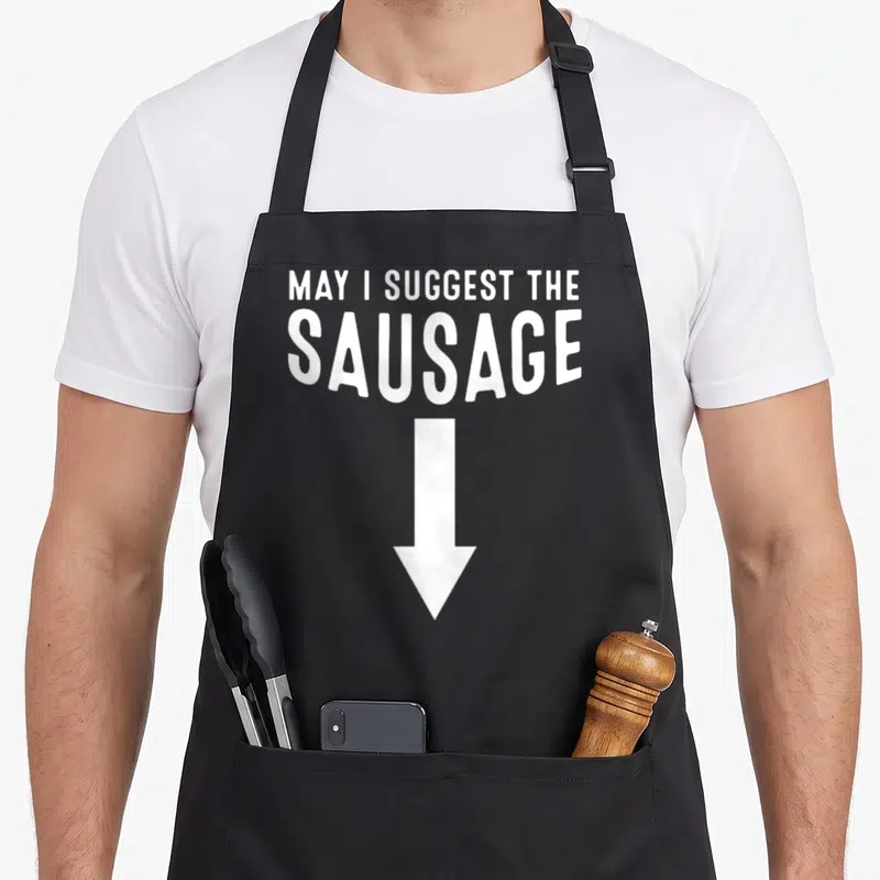 Inappropriate sausage apron - White text MAY I SUGGEST THE SAUSAGE with downward arrow — shown on a black cotton canvas apron against a white background