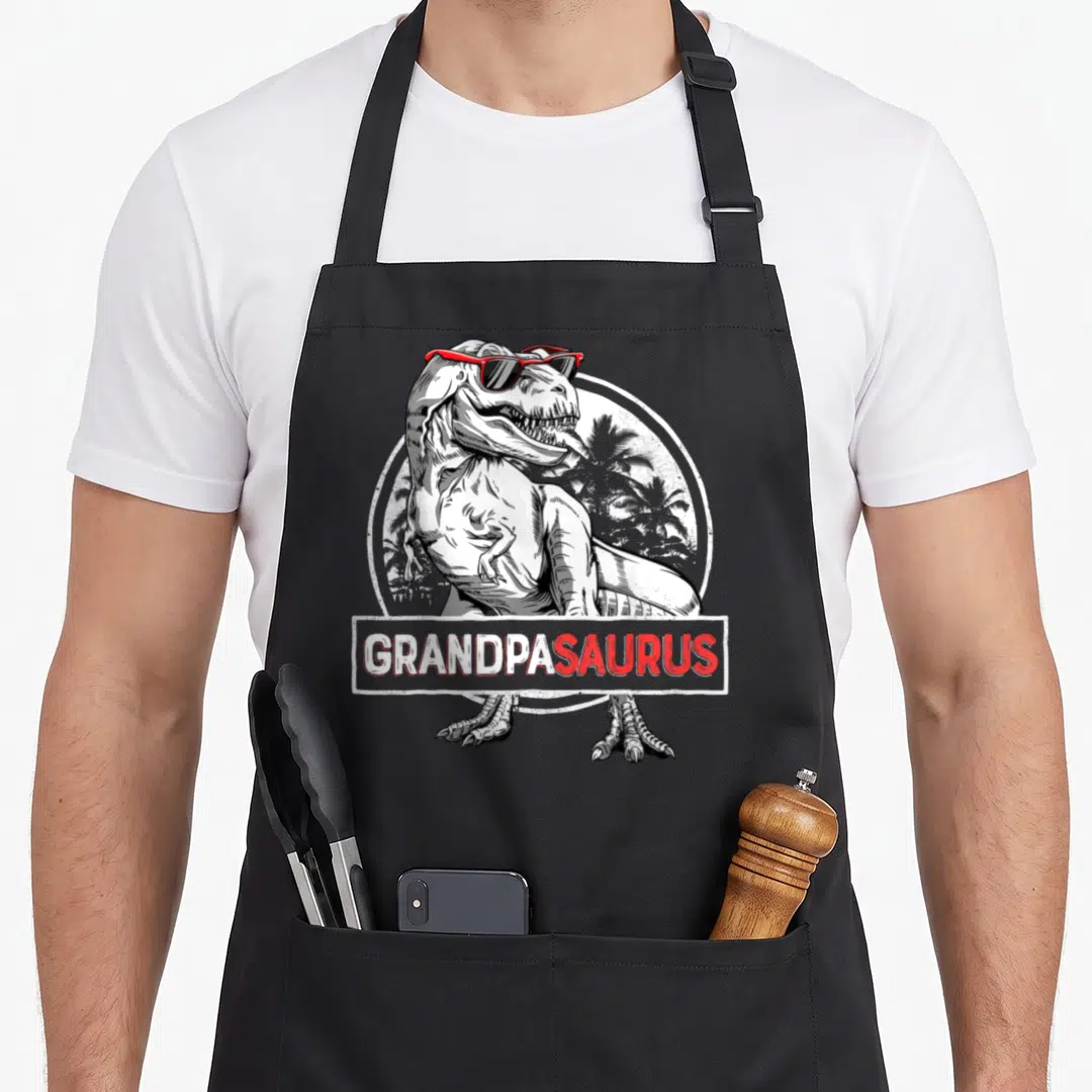 Grandpa t-rex apron - T-Rex wearing red sunglasses with Grandpasaursus text banner and palm tree circle background — shown on a black cotton canvas apron against a white background