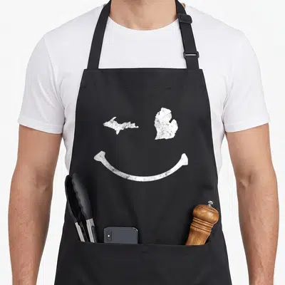 Winking michigan apron - White distressed smiley face formed by two Michigan island maps and a curved smile line — shown on a black cotton canvas apron against a white background
