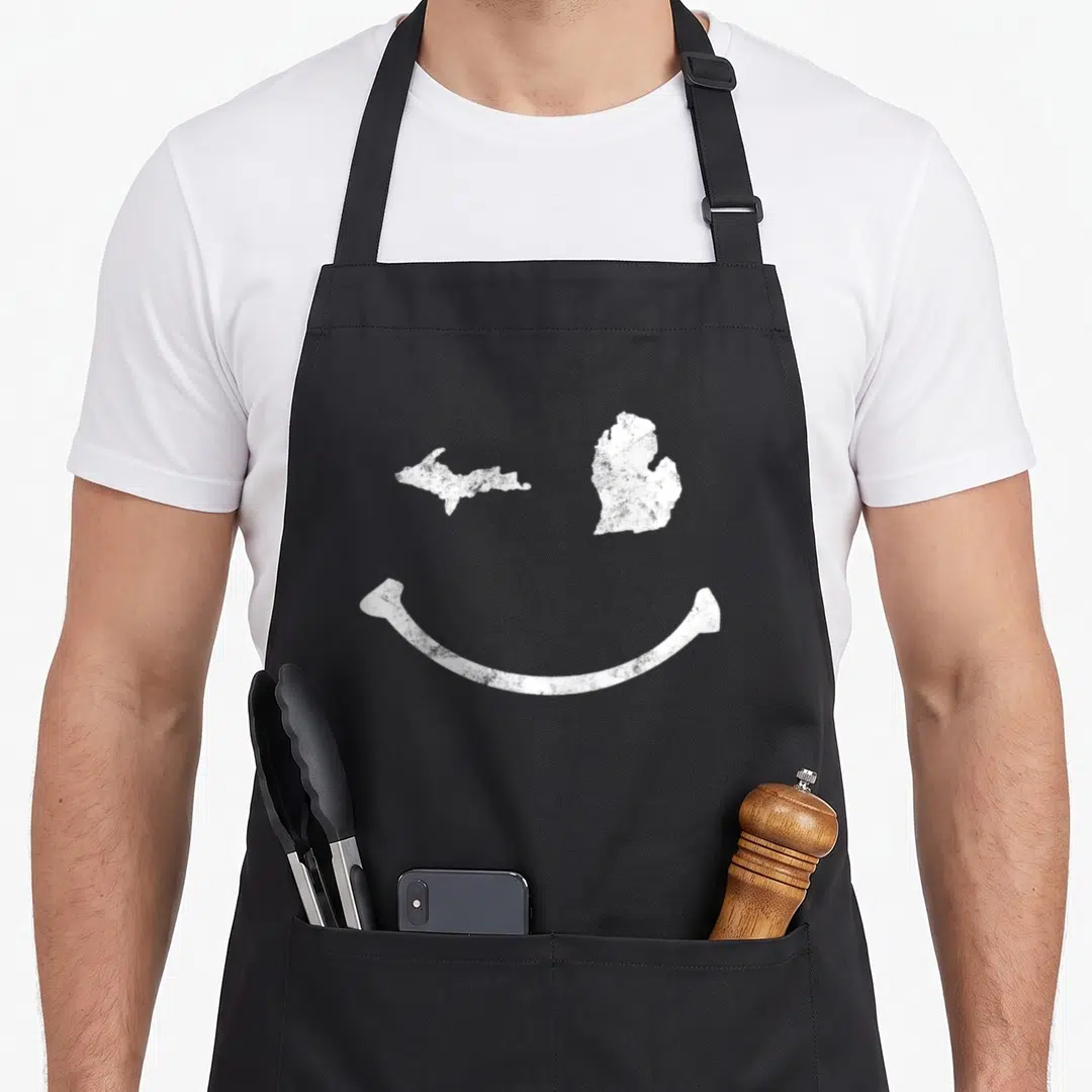 Winking michigan apron - White distressed smiley face formed by two Michigan island maps and a curved smile line — shown on a black cotton canvas apron against a white background