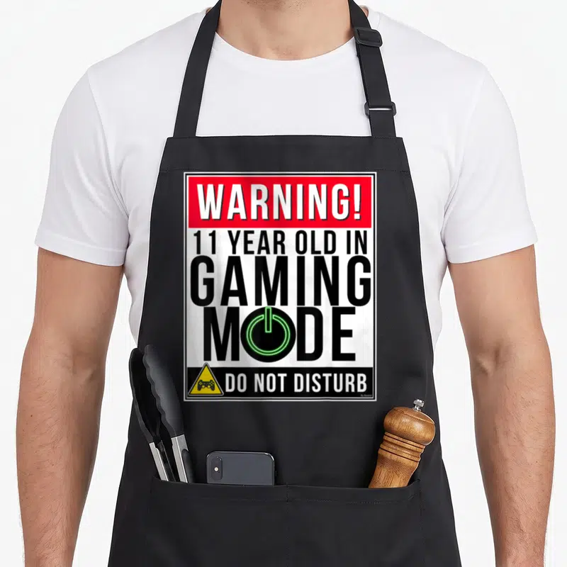 11th gamer birthday apron - Rectangular warning sign print with red header, green power symbol in Gaming Mode text — shown on a black cotton canvas apron against a white background