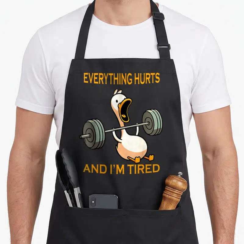Gym duck apron - Cartoon goose lifting a barbell with orange text reading Everything Hurts And I'm Tired on a grey — shown on a black cotton canvas apron against a white background