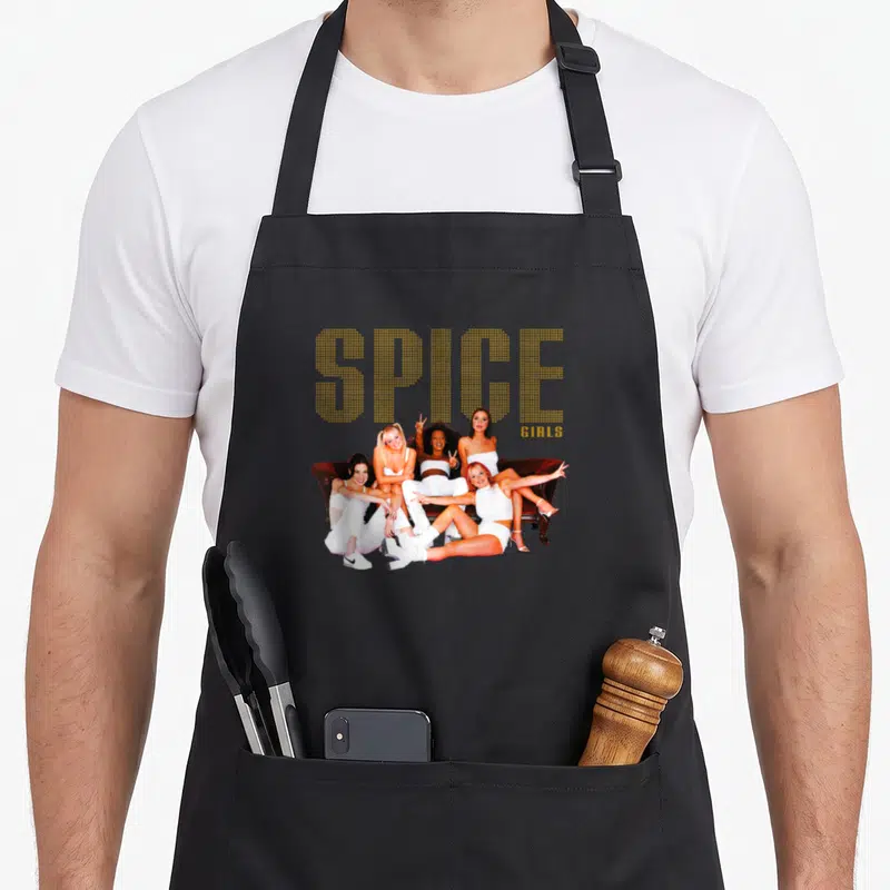 Couch spice apron - Gold SPICE GIRLS text above five women in white posing on a brown sofa — shown on a black cotton canvas apron against a white background