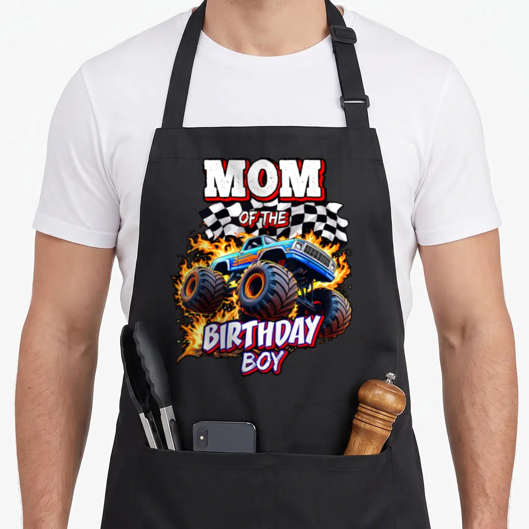 Cool birthday truck apron - Monster truck graphic with fire and MOM OF THE BIRTHDAY BOY text in cartoon style — shown on a black cotton canvas apron against a white background