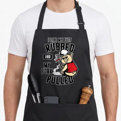 Ich meat aprons - Cartoon pig in red vest holding sausage with text I like my butt rubbed and my pork pulled — shown on a black cotton canvas apron against a white background