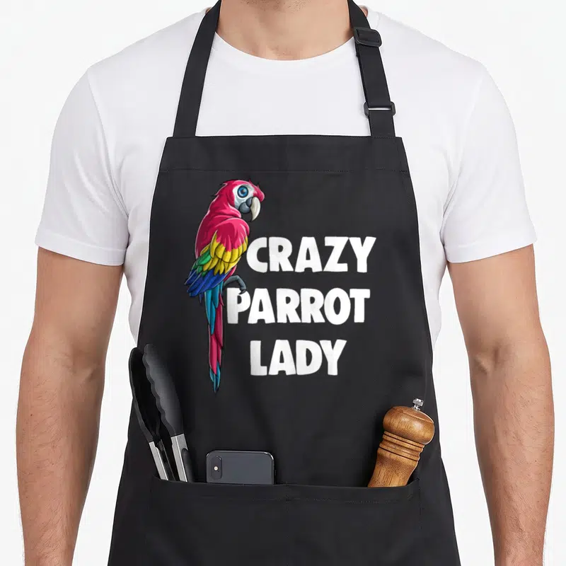 Cute crazy parrot lady apron - Cartoon parrot with pink, yellow, blue feathers beside stacked white text reading CRAZY — shown on a black cotton canvas apron against a white background