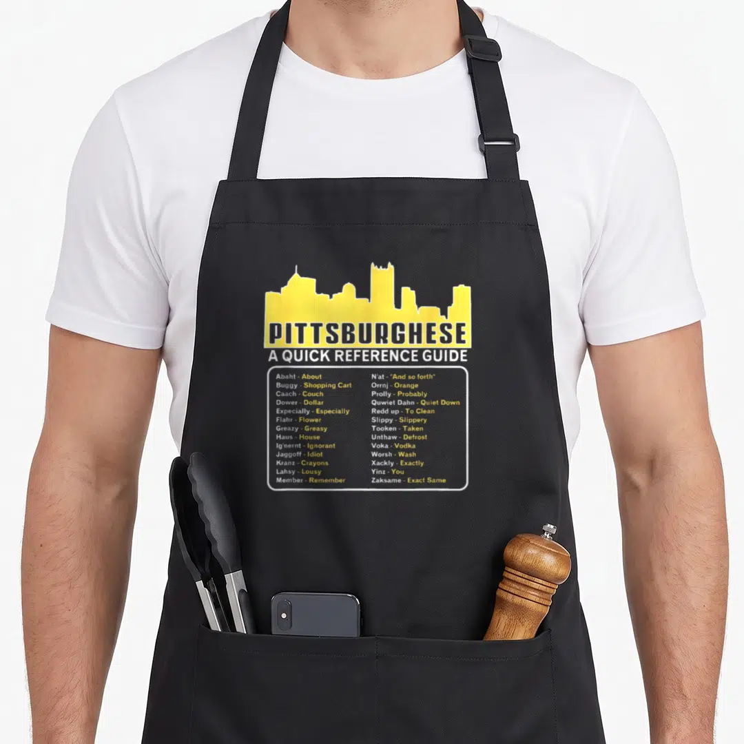 Pittsburghese quick apron - Yellow Pittsburgh skyline above two columns of dialect words like Haus and Voka in white — shown on a black cotton canvas apron against a white background