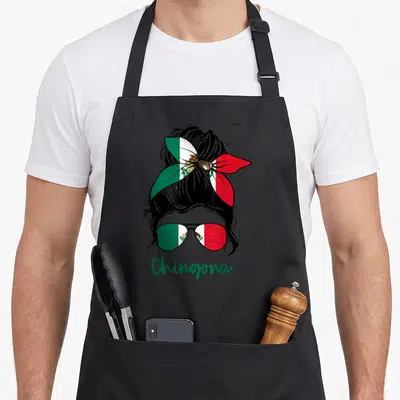 Chingona mexican apron - Cartoon woman head with bun and sunglasses filled with Mexican flag pattern and green text — shown on a black cotton canvas apron against a white background