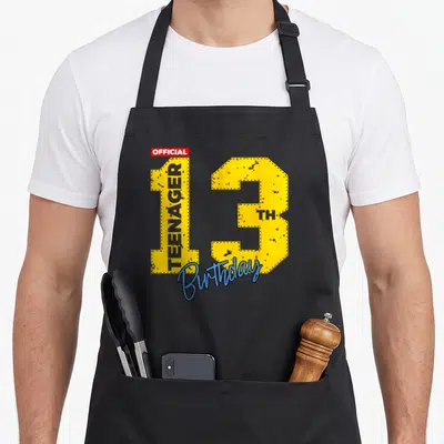 Funny gamer birthday apron - Yellow number 13 with official teenager text and blue birthday script on red accent — shown on a black cotton canvas apron against a white background
