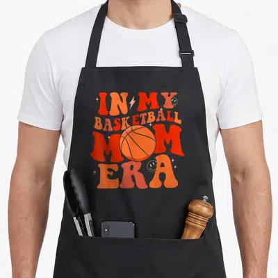 Basketball mom era apron - Retro orange and red text reading In My Basketball Mom Era with a central basketball — shown on a black cotton canvas apron against a white background