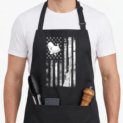 Turkey grill hunter apron - White distressed flag with turkey silhouette in stars and shotgun in stripes — shown on a black cotton canvas apron against a white background