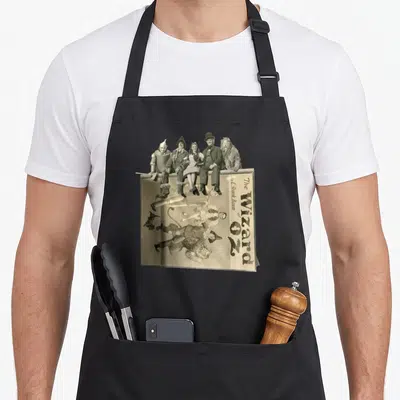 Tin dorothy apron - Five characters sit on inverted Wizard of Oz book cover text in sepia tones — shown on a black cotton canvas apron against a white background