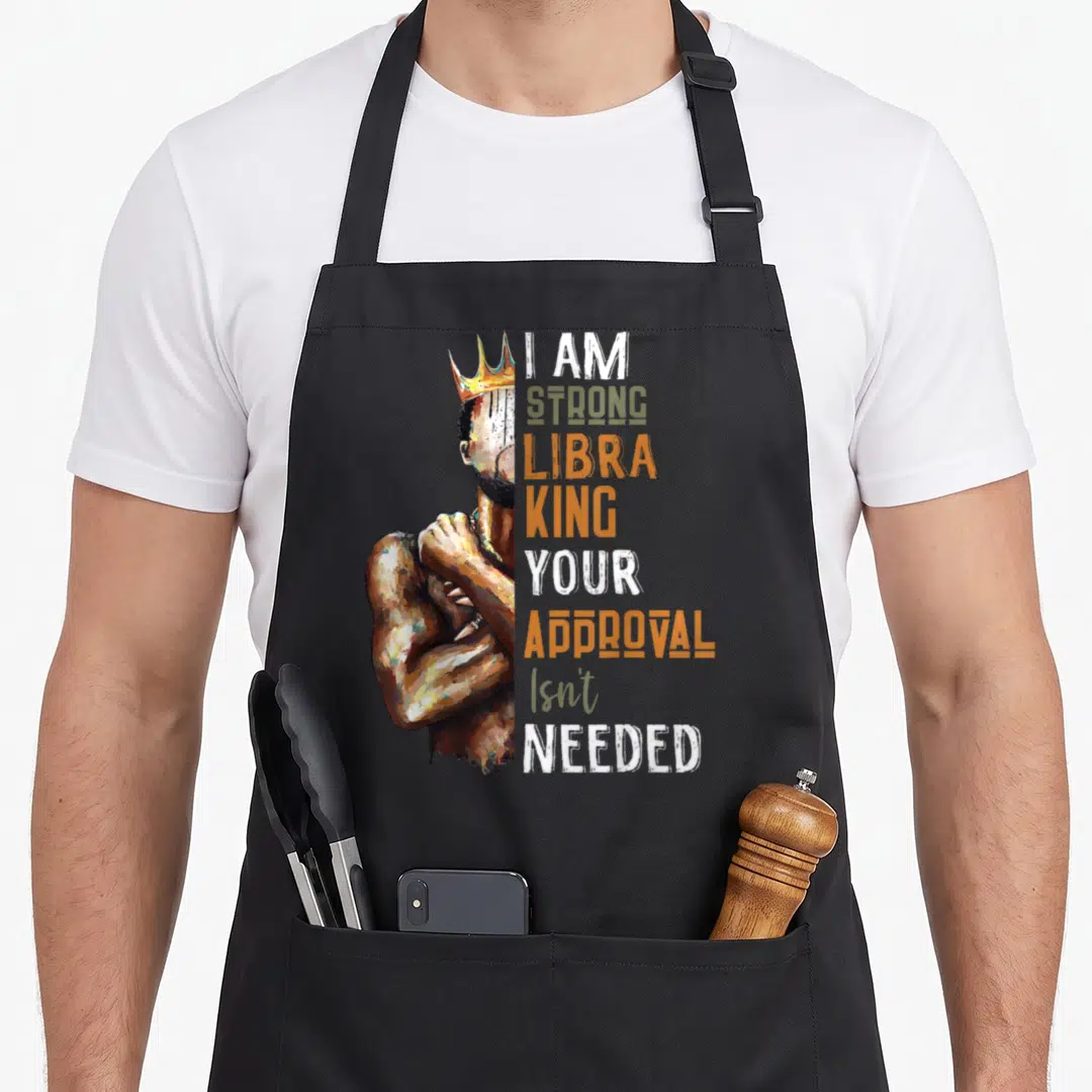 Strong king apron - Crowned man flexing arm next to I AM STRONG LIBRA KING text — shown on a black cotton canvas apron against a white background