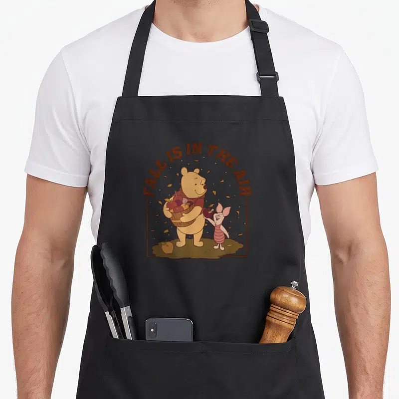Air thanksgiving apron - Winnie the Pooh holding red apples under brown text with Piglet on brown ground — shown on a black cotton canvas apron against a white background