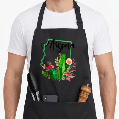 State map cactus pride apron - Arizona script over green state outline with watercolor cacti and pink orange flowers — shown on a black cotton canvas apron against a white background