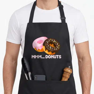 Mmm donut apron - Two cartoon doughnuts pink and chocolate with mmm donuts text — shown on a black cotton canvas apron against a white background
