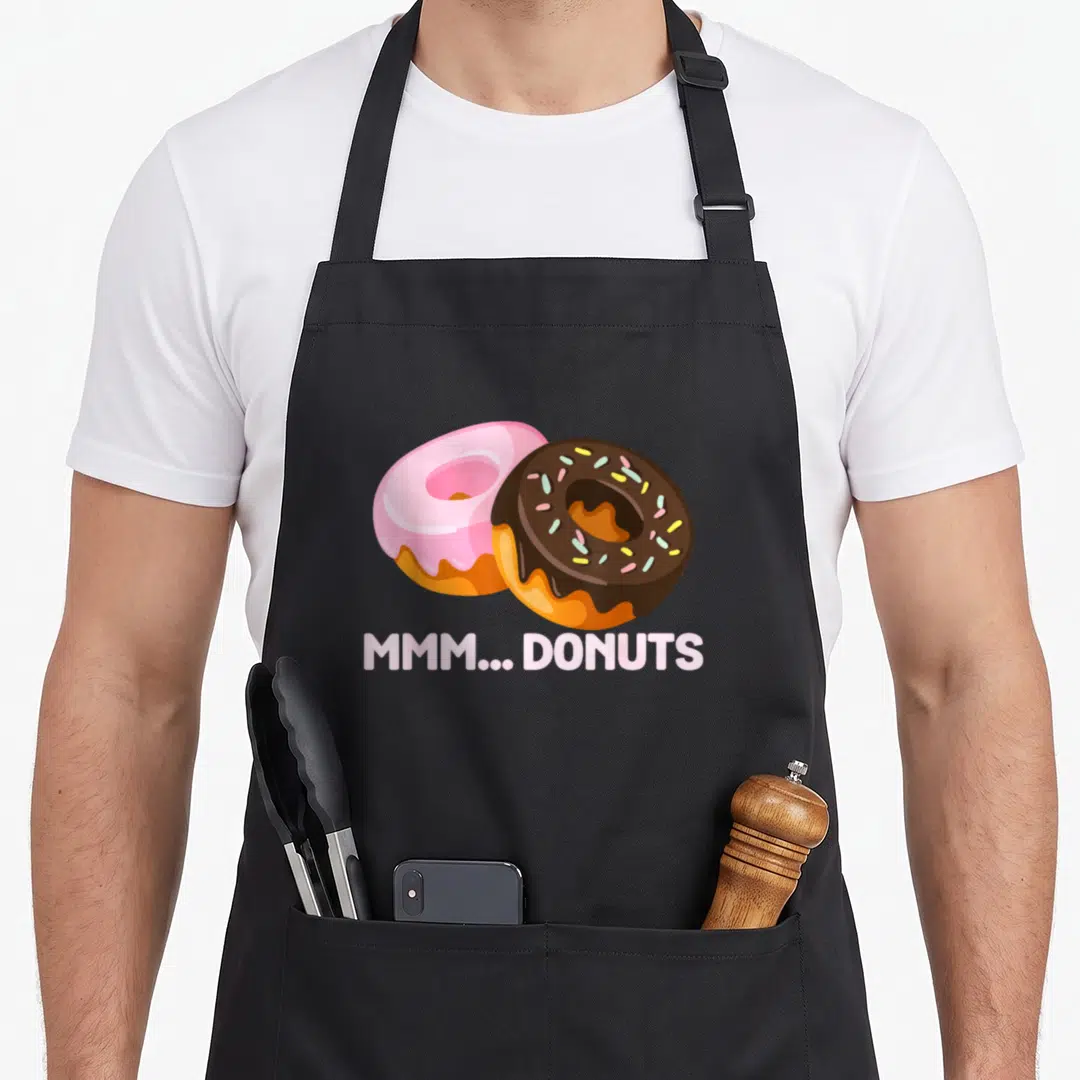 Mmm donut apron - Two cartoon doughnuts pink and chocolate with mmm donuts text — shown on a black cotton canvas apron against a white background