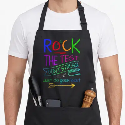 Testing test kitchen apron - Stacked colorful text ROCK THE TEST with leaf, ribbon, paper plane and arrow graphics — shown on a black cotton canvas apron against a white background