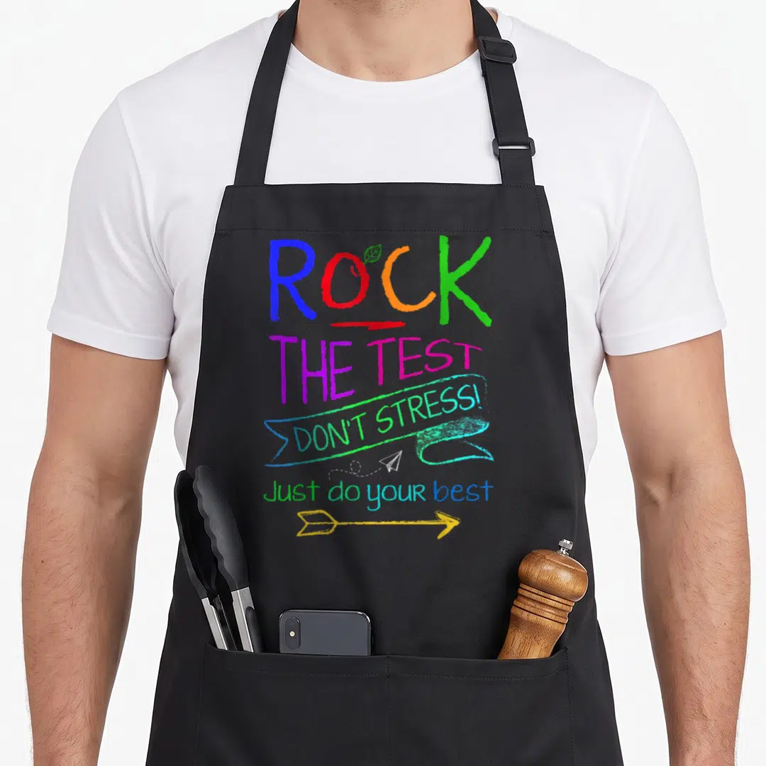 Testing test kitchen apron - Stacked colorful text ROCK THE TEST with leaf, ribbon, paper plane and arrow graphics — shown on a black cotton canvas apron against a white background