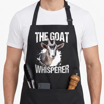 Whisperer goat apron - Realistic goat head between arched The Goat Whisperer text over white splatter — shown on a black cotton canvas apron against a white background