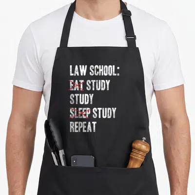 Funny law student aprons - LAW SCHOOL text with red Xs crossing out EAT and SLEEP — shown on a black cotton canvas apron against a white background