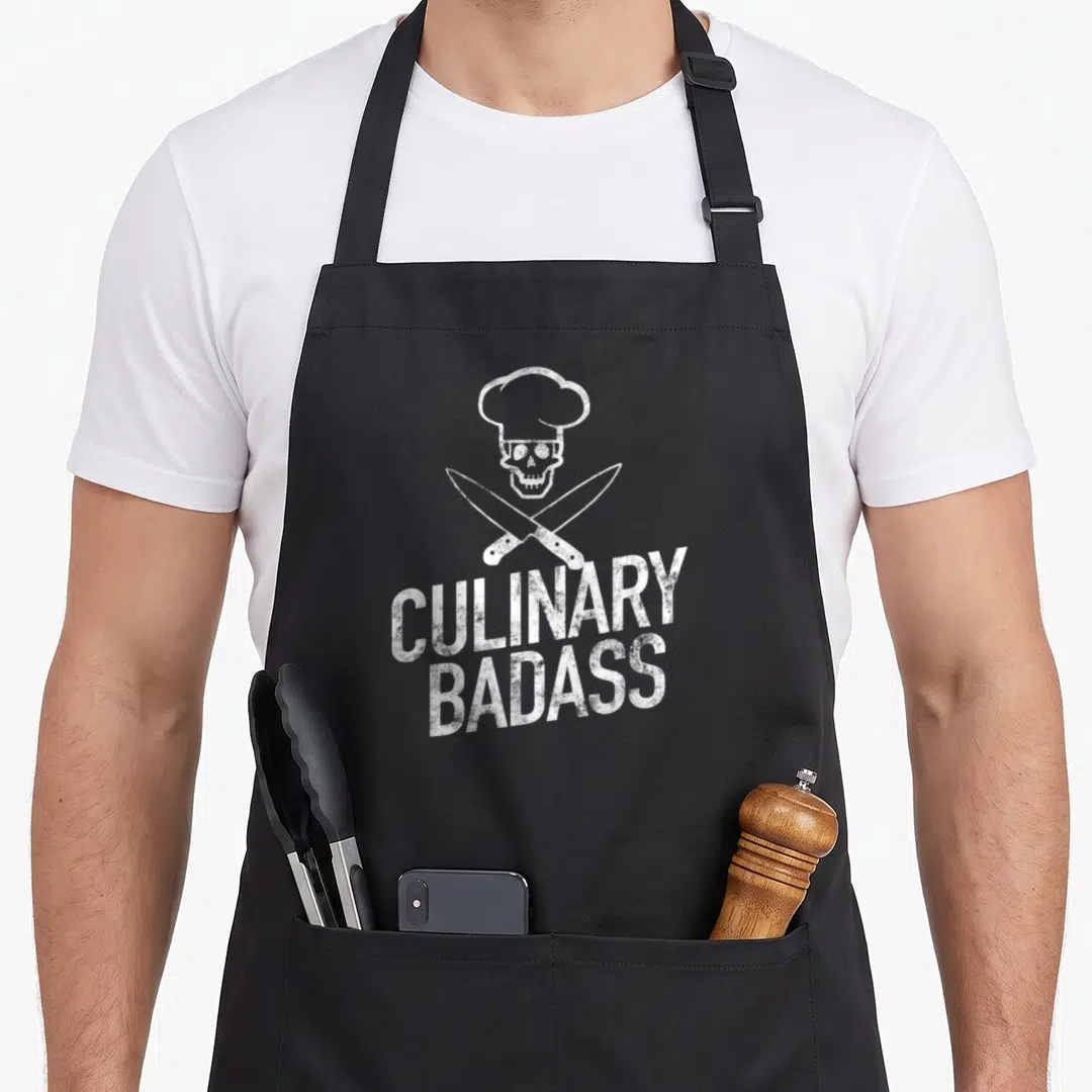 Funny culinary badass apron - White distressed skull in chef hat above crossed knives with CULINARY BADASS text — shown on a black cotton canvas apron against a white background