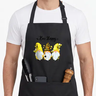 Bee spring apron - Three gnomes with bee-striped hats and Bee Happy text in yellow and black — shown on a black cotton canvas apron against a white background