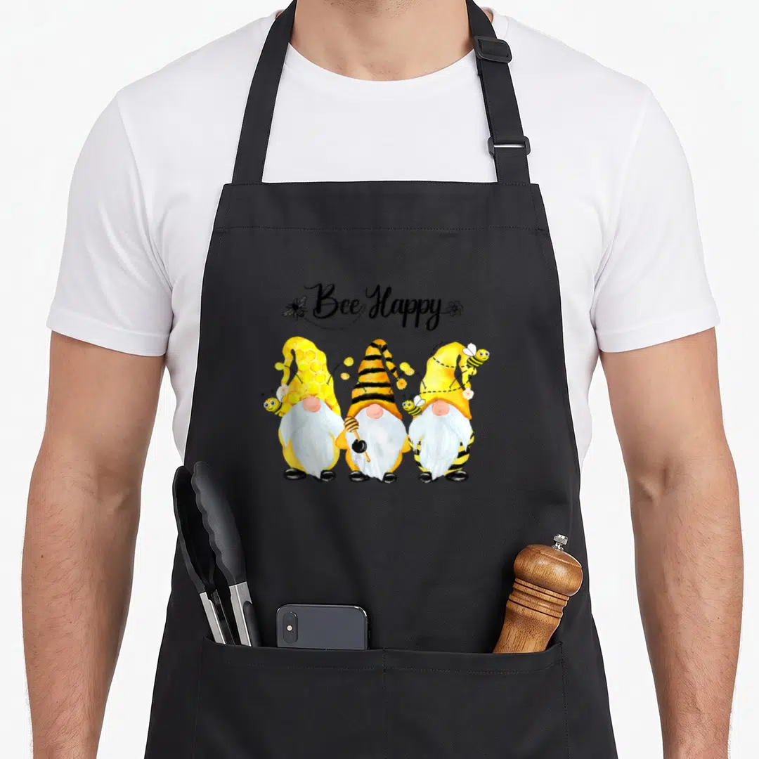 Bee spring apron - Three gnomes with bee-striped hats and Bee Happy text in yellow and black — shown on a black cotton canvas apron against a white background