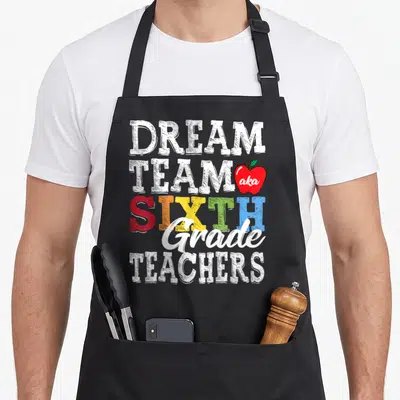 Sixth cute teacher aprons - Stacked text DREAM TEAM SIXTH Grade TEACHERS with red apple containing aka — shown on a black cotton canvas apron against a white background