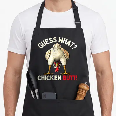 Funny butt chicken apron - Cartoon chicken illustration with GUESS WHAT? CHICKEN BUTT! text in white and red — shown on a black cotton canvas apron against a white background
