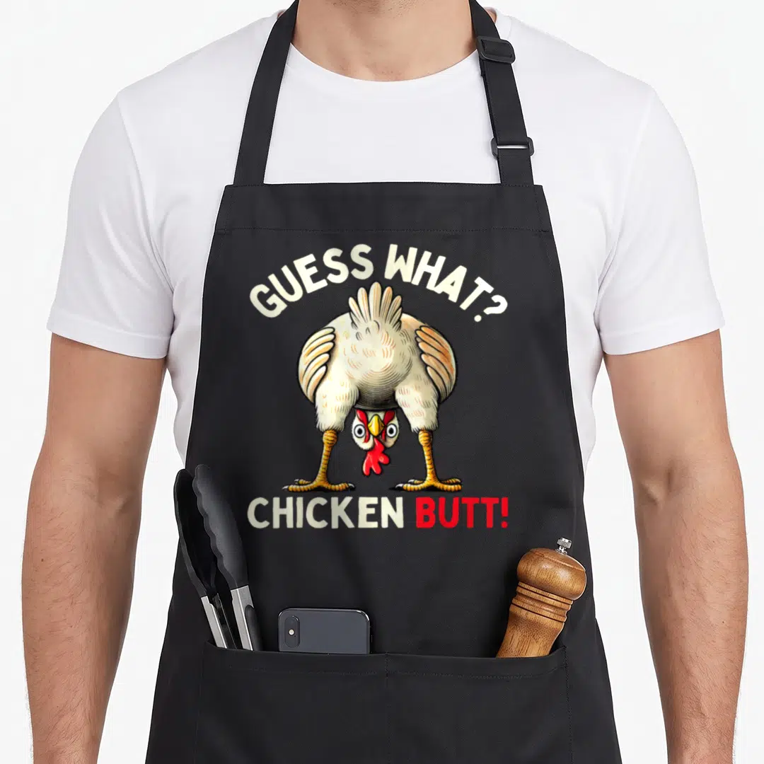 Funny butt chicken apron - Cartoon chicken illustration with GUESS WHAT? CHICKEN BUTT! text in white and red — shown on a black cotton canvas apron against a white background