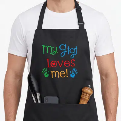 Gigi grandchild apron - Stacked text my Gigi Loves me! with red heart in O and green blue handprints — shown on a black cotton canvas apron against a white background