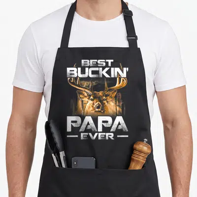 Buckin papa apron - White and orange distressed deer head with text BEST BUCKIN PAPA EVER flanked by horizontal lines — shown on a black cotton canvas apron against a white background