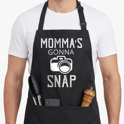 Mommas snap aprons - White distressed text Momma's Gonna Snap with camera icon — shown on a black cotton canvas apron against a white background